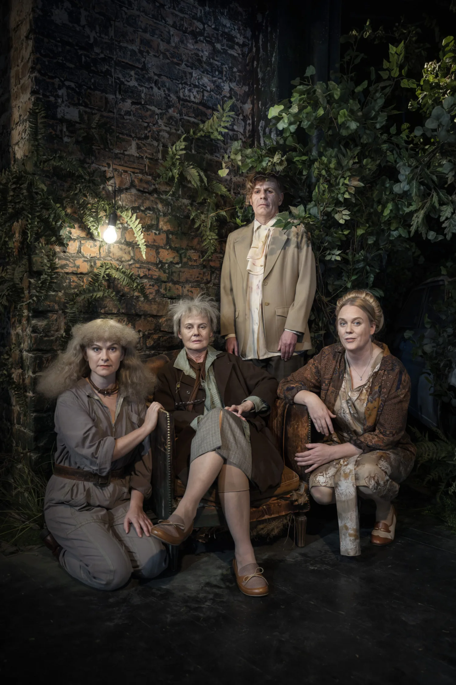 Three people sitting and one standing in the dark bby a brick wall with greenry and a small street light.