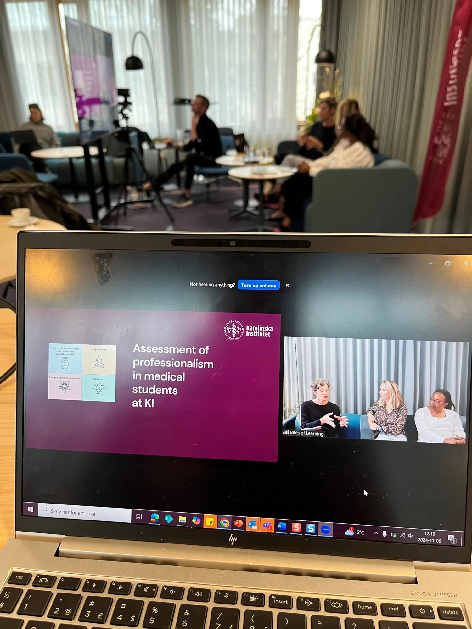 A computers screen with a webinar on, in background is the presenters sitting in a sofa and discuss.