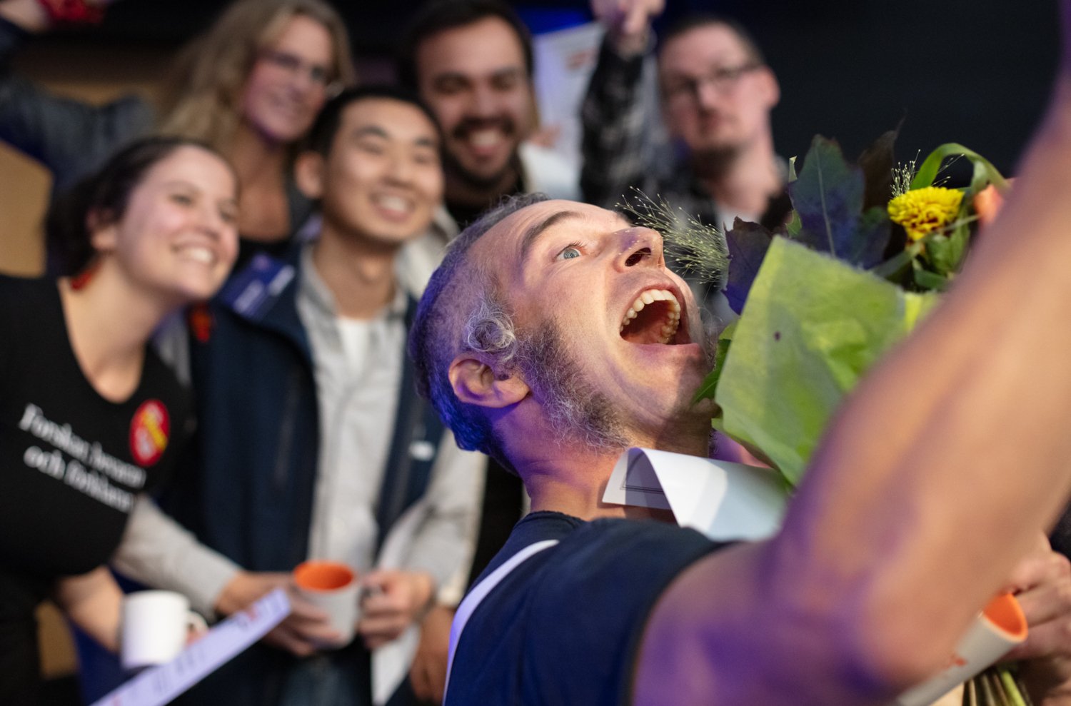Happy man taking a selfie together with five people in the background whose faces are a bit blurred.