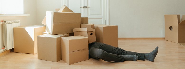 Genre image of a man laying under several moving boxes.
