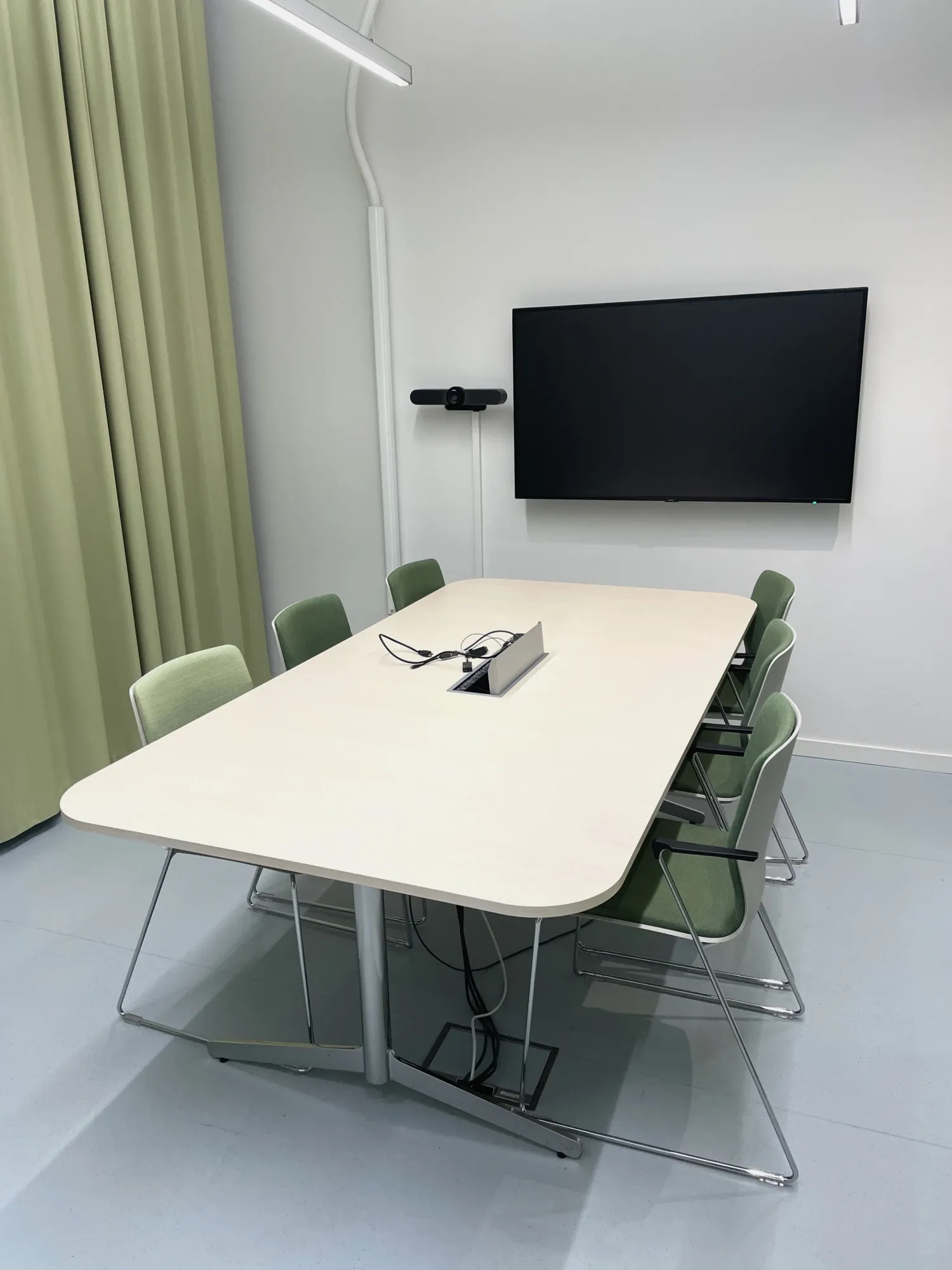 A meeting room with a square table with six chairs around it. On the wall is a TV screen and a webcam.