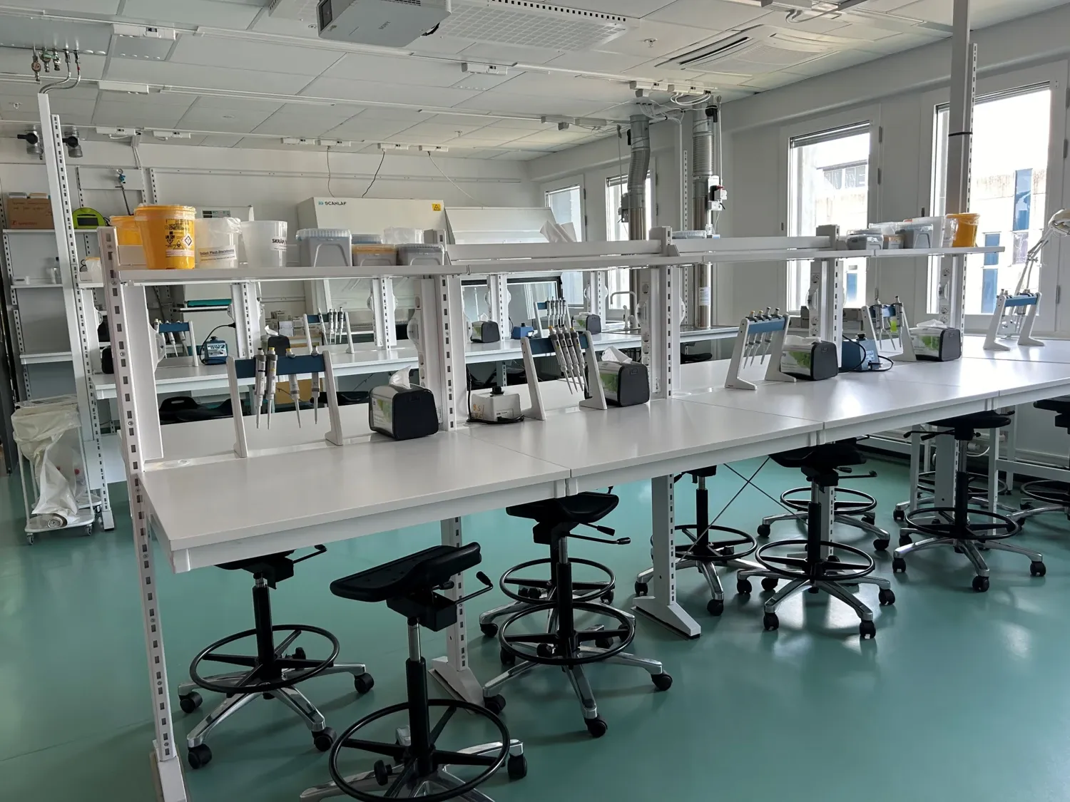 Photo of a lab with white laboratory benches, chairs and a green floor.