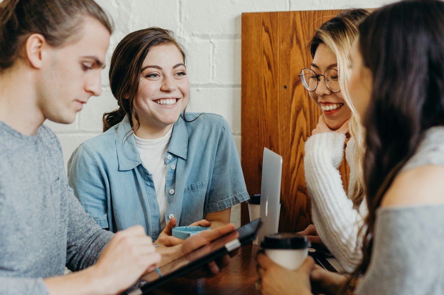 A group of young adults sitting aroud a table smiling, one of them has a coffee mug in their hand