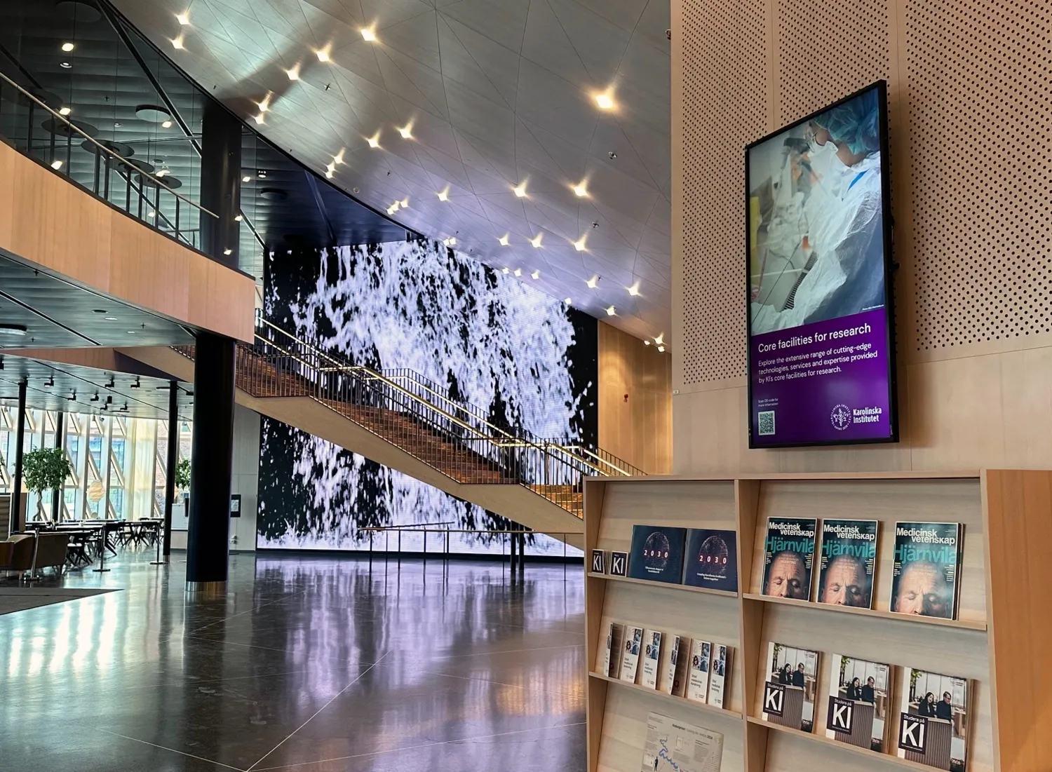 Digital screen next to reception above shelf with magazines and other printed material. In the background you can see slideshows on the wall and the stairs up to level 3.