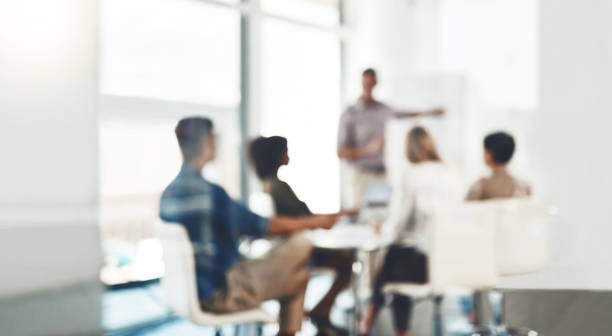 Blurry image of people sitting in a meeting. Four are sitting down and one standing, pointing at a board,