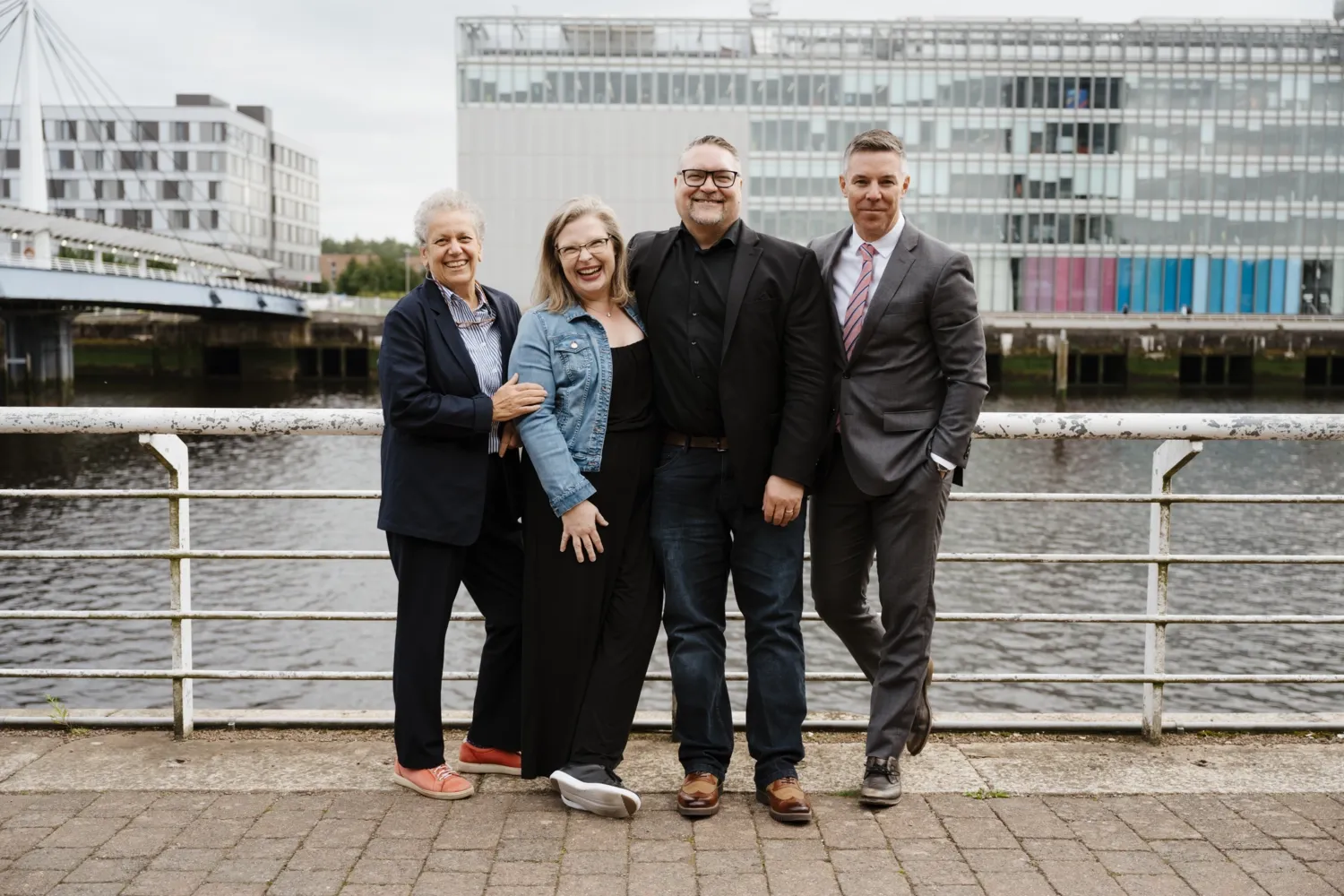 The four hosts of the Paper poscast stand by the quayside, large buildings loom in the background. They stand close to each other and look happy.