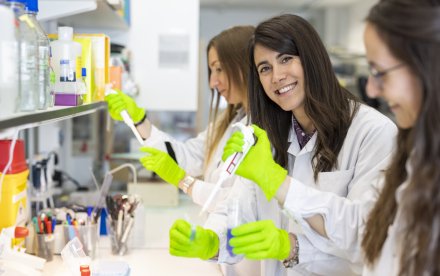 Three femare researchers standing with test tubes and pippettes in lab environment. One of them is looking in to the camera.