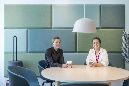 Two women sit at a table with coffee in their hands and look into the camera.