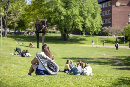 Woman with badminton racket bag sitting on her back on lawn on campus Solna, with people in the background.